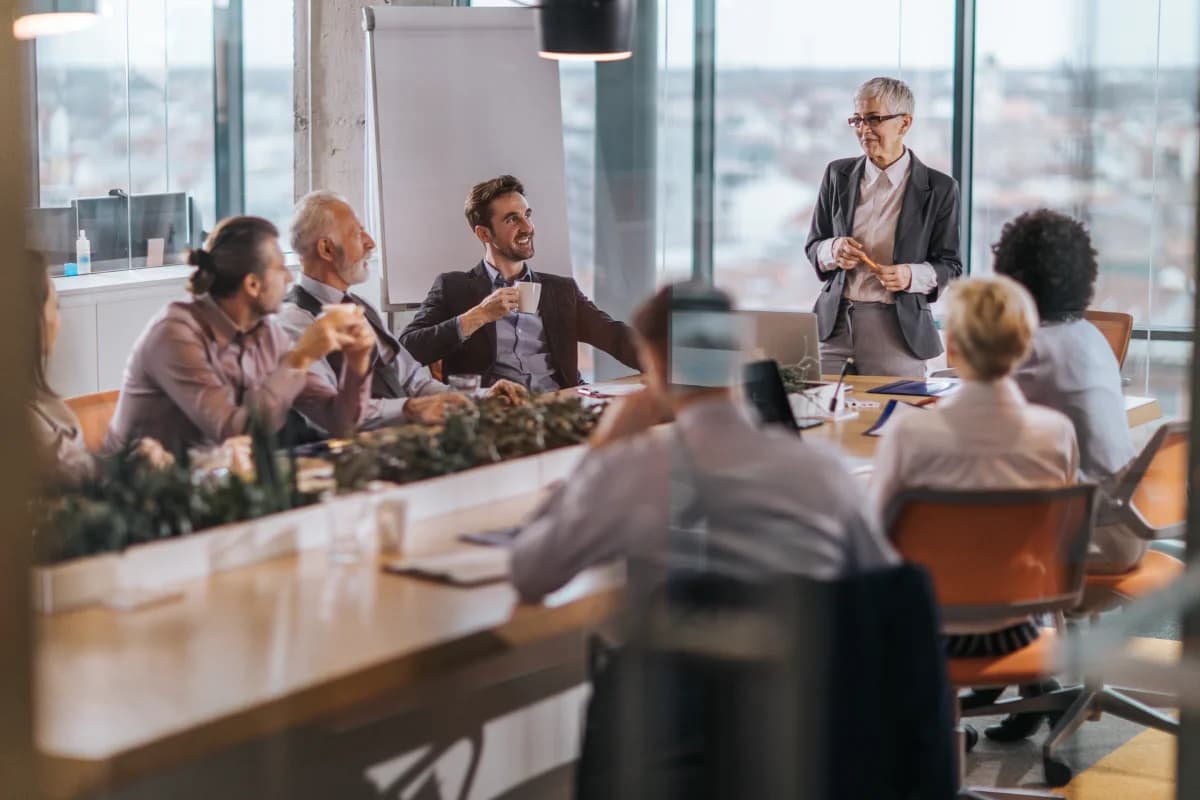 A group of professionals sit around a conference table in a modern office. One person stands and speaks while others listen, with notebooks and papers on the table and large windows overlooking a city in the background.